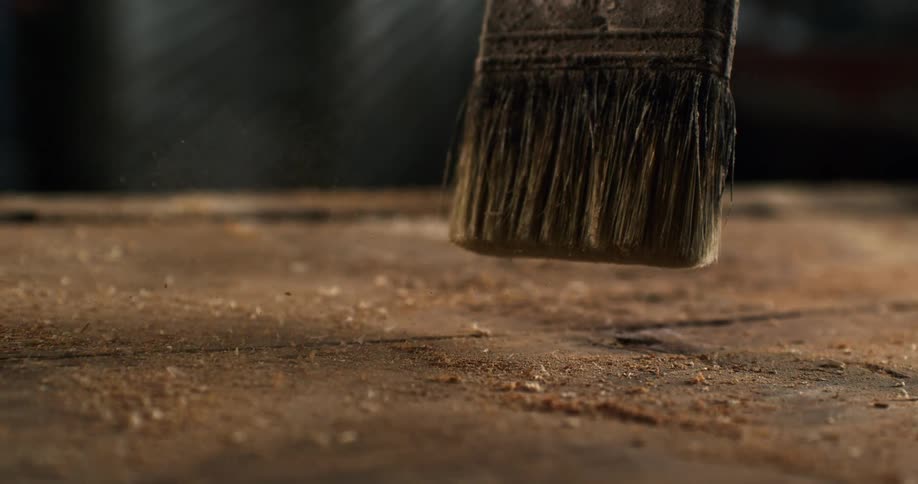 A paintbrush sweeps sawdust and dust off of a rustic wooden workbench in a workshop in super slow motion