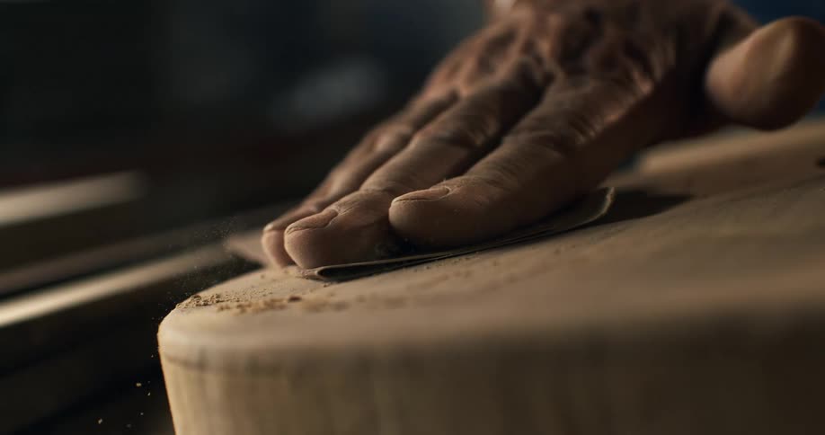 A craftsman's experienced hand meticulously sands a piece of wood in a workshop, with fine dust flying in slow motion