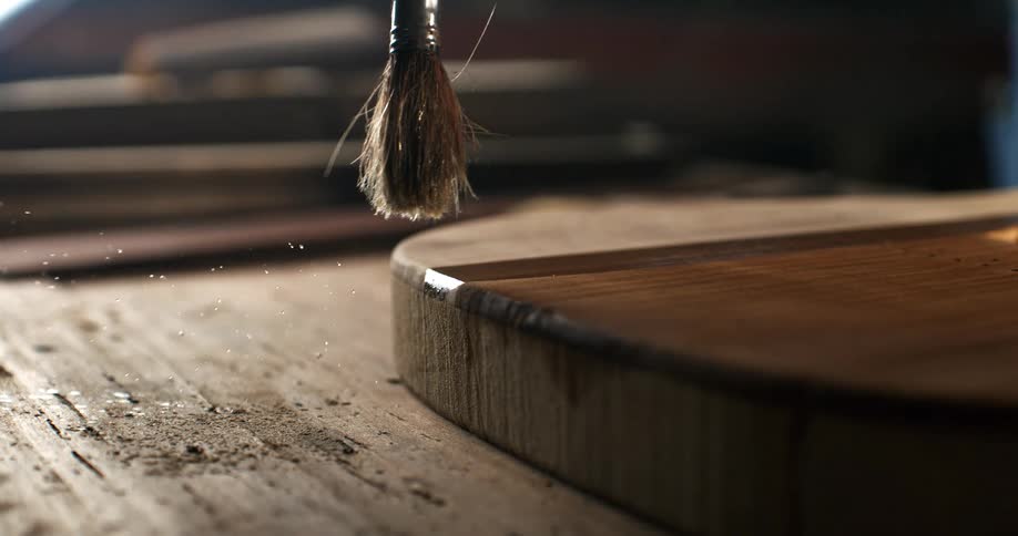 Extreme slow motion close up of a paintbrush applying varnish to a wooden board in a dusty workshop