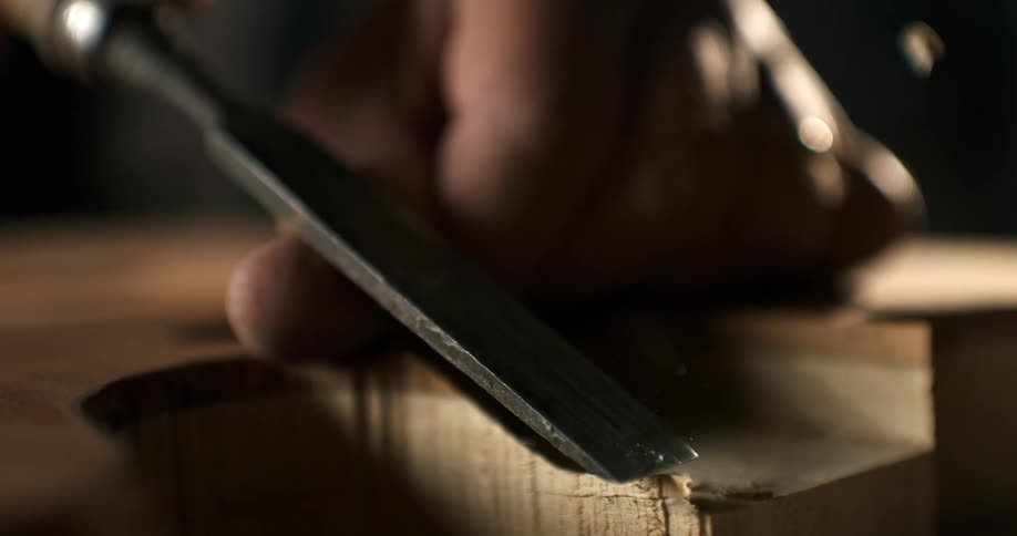 An artisan woodworker carves a piece of lumber with a chisel, creating a delicate wood shaving in dramatic slow motion
