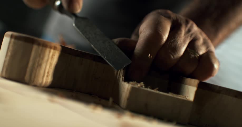 A luthier's hands meticulously carve a wooden instrument with a chisel in dramatic slow motion