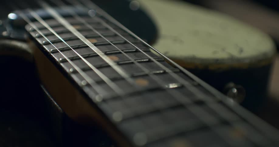 A slow, close-up dolly shot moves down the fretboard of a weathered electric guitar, highlighting the strings and frets