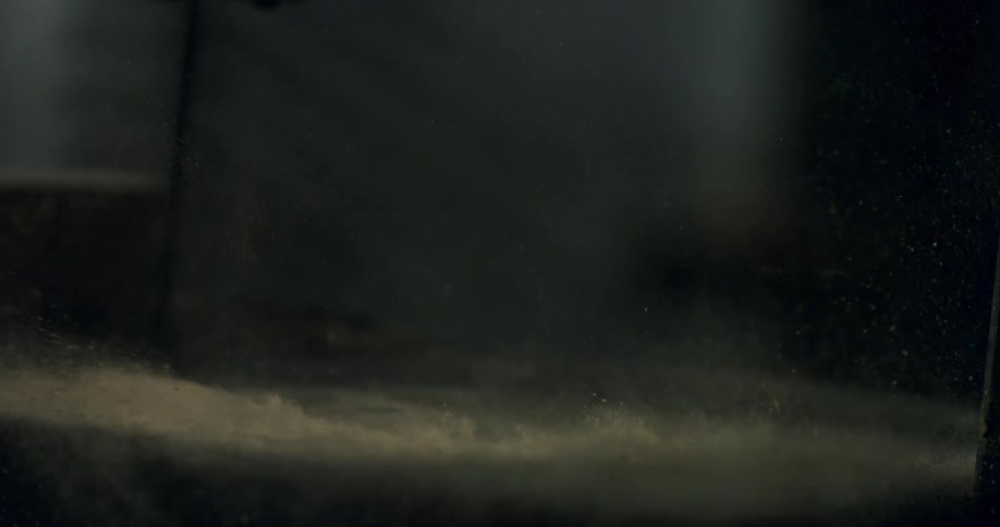 A gust of wind blows a cloud of dust and particles across a dark floor in dramatic slow motion