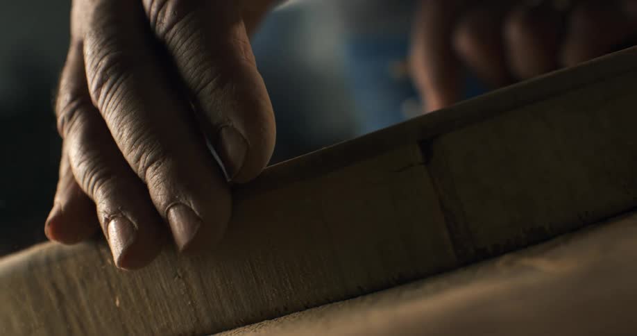 In slow motion, a carpenter's hands wipe away fine sawdust from a piece of carved wood in a dimly lit workshop
