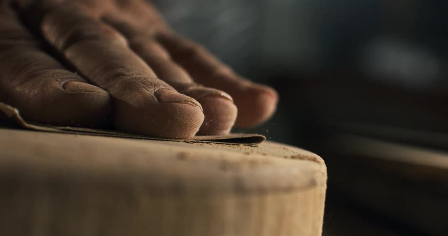 A woodworker's hand meticulously sands the smooth edge of a circular wooden piece in dramatic slow motion
