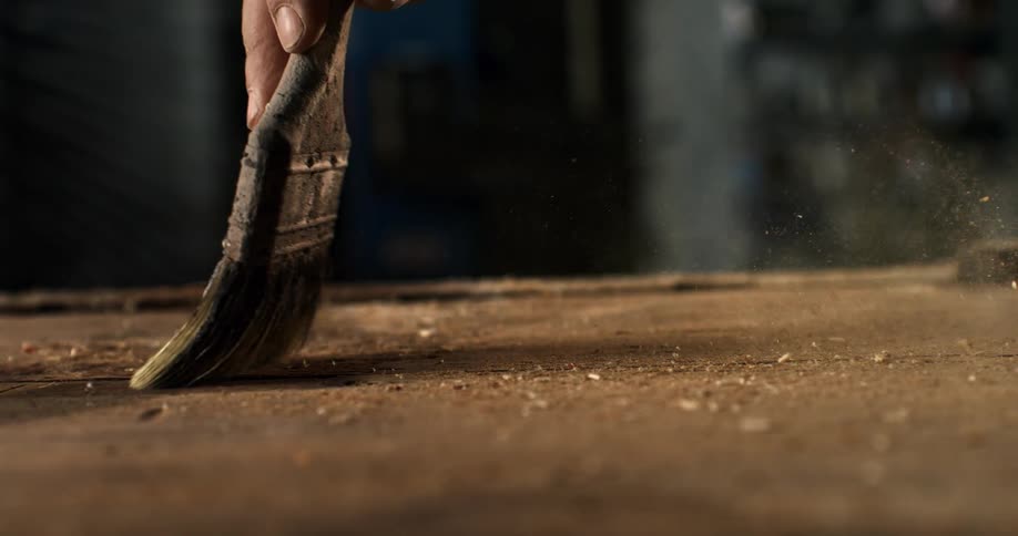 A carpenter's hand sweeps sawdust from a wooden workbench with an old paintbrush in slow motion