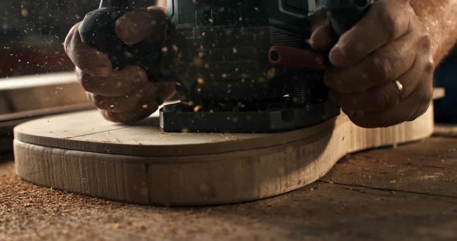 A craftsman carefully uses a router to shape the wooden body of a custom guitar in slow motion, sending sawdust flying