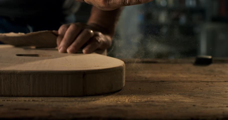 A luthier meticulously sands the wooden body of a handmade guitar, with sawdust dancing in the air in slow motion