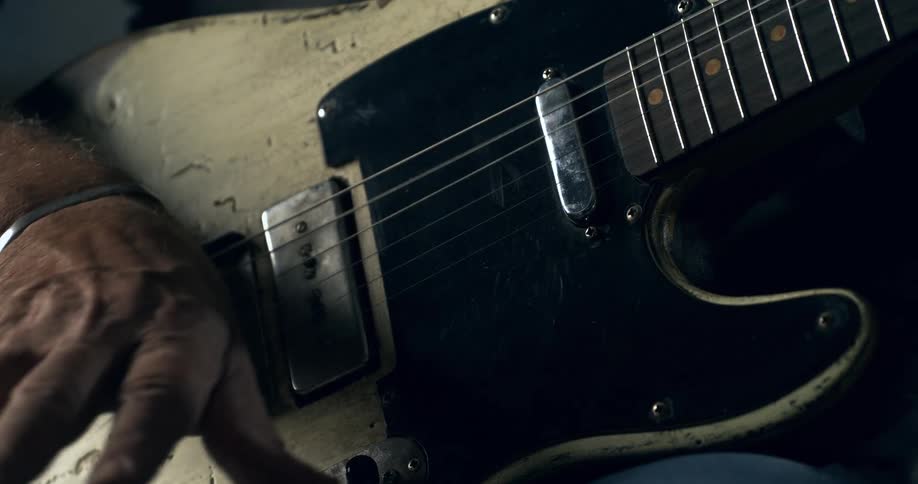 A musician strums and palm-mutes the strings of a well-worn, vintage-style electric guitar in a moody, close-up shot