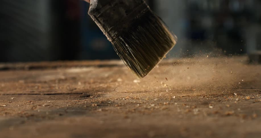 A well-worn paintbrush sweeps sawdust from a wooden workbench in a dramatic slow-motion close-up