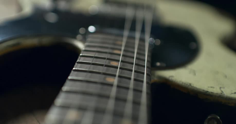 A slow, shallow-focus shot moving along the strings and fretboard of a distressed, vintage-style electric guitar