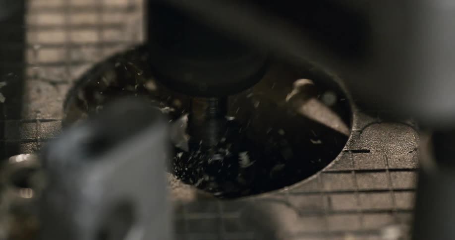 An overhead slow-motion macro shot shows a CNC milling machine carving a circular hole, sending debris flying