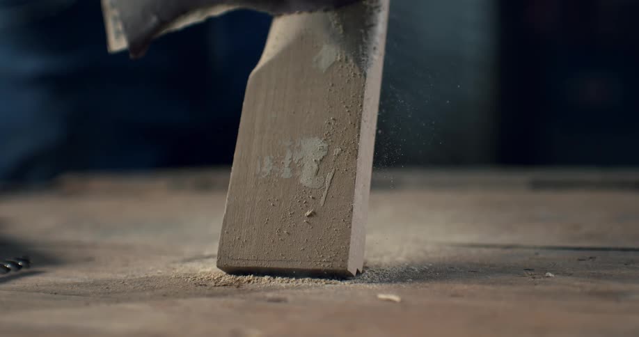 A craftsman uses sandpaper on a wooden block in slow motion, with sawdust flying off