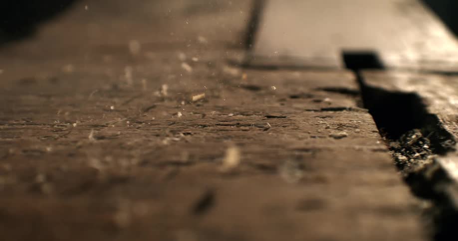 Slow motion macro shot of dust and wood shavings rising from a weathered wooden surface in a dimly lit room