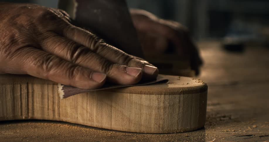 Skilled hands of a luthier meticulously sand the body of a custom guitar in a workshop, with sawdust flying in slow motion