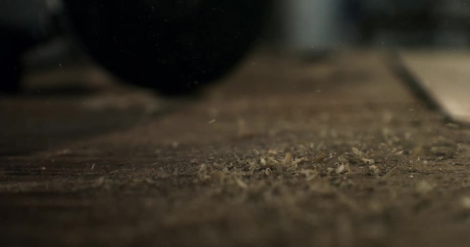 A dramatic slow-motion close-up captures sawdust and wood shavings being blown across a wooden surface