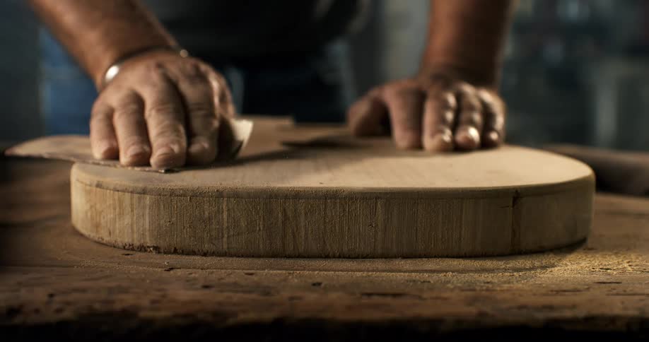 A luthier's hands carefully sand the wooden body of a custom guitar in a dusty workshop in slow motion