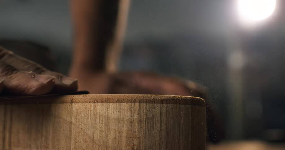 A craftsman's experienced hand carefully sands a wooden block, with sawdust particles flying into the air in cinematic super slow motion