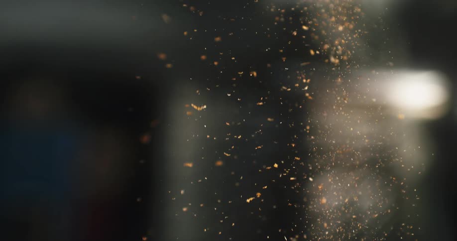 Abstract slow-motion close-up of golden sawdust particles floating and falling through the air, backlit by a soft light