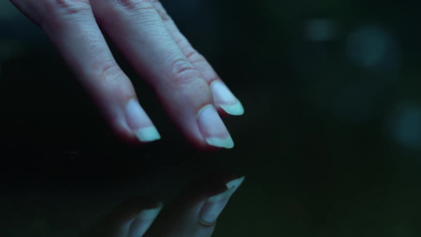Close-up of a woman's hand with long natural nails gently hovering over still, dark water, creating a mysterious reflection