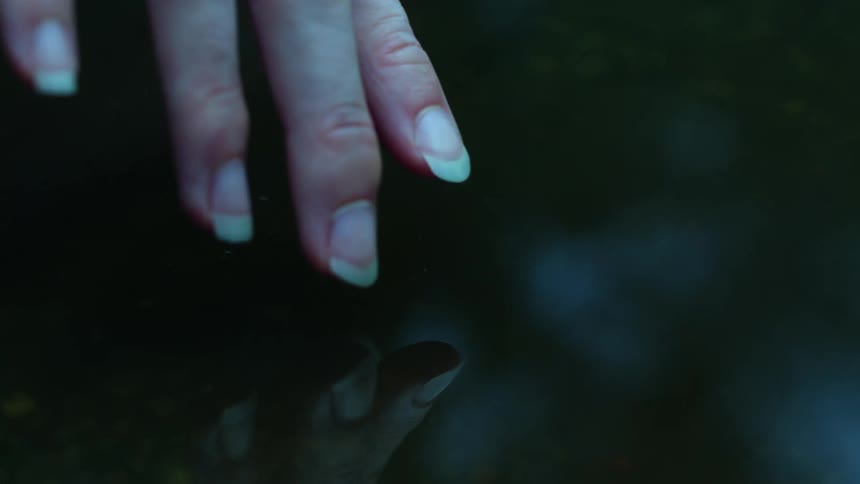 A woman's hand hovers just above the surface of dark, still water, creating a reflection