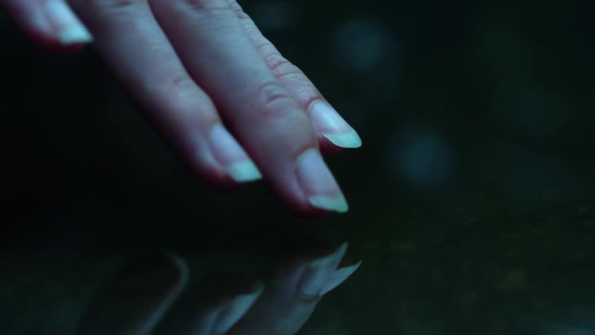 A woman's fingers gently touch the surface of still, dark water in slow motion, creating expanding ripples