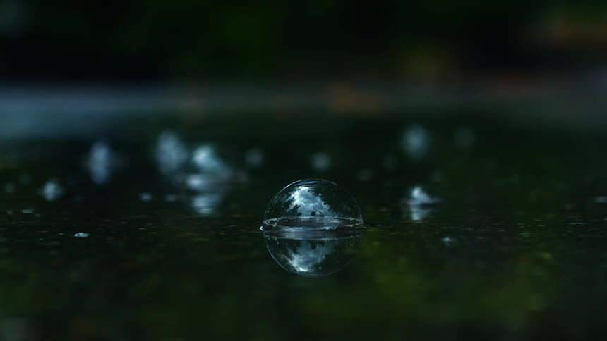 A single bubble reflecting the sky floats on a dark puddle before popping in dramatic slow motion