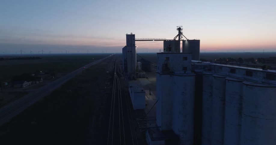 An aerial drone flies towards a large grain elevator in a rural American town at dusk, revealing a highway, railroad tracks, and wind turbines on the horizon