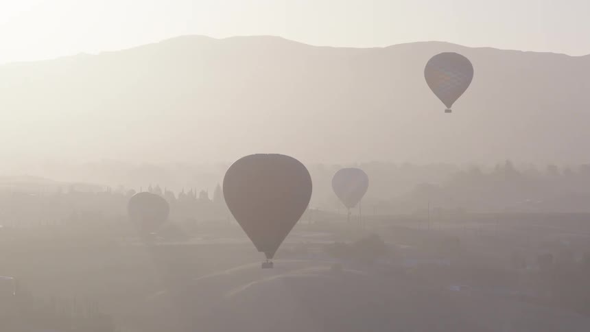 Silhouetted hot air balloons float over a hazy valley as one descends to land on a rolling hill at sunrise