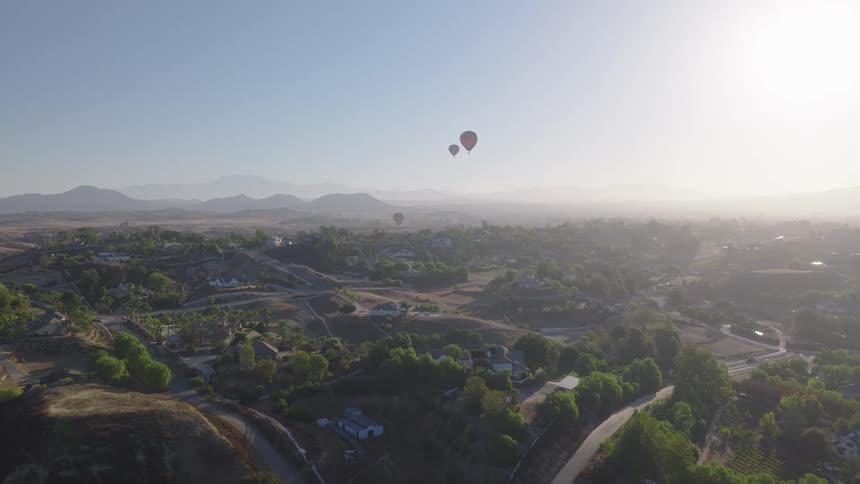 Hot air balloons float gracefully over a lush, green valley town during a hazy sunrise, with distant mountains on the horizon