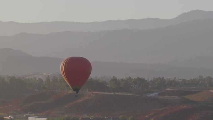 A red hot air balloon floats peacefully over rolling hills with hazy mountains in the background during a serene sunrise