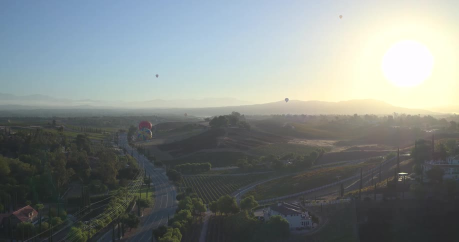 A breathtaking aerial view shows hot air balloons gliding over the rolling hills and vineyards of Temecula wine country at sunrise