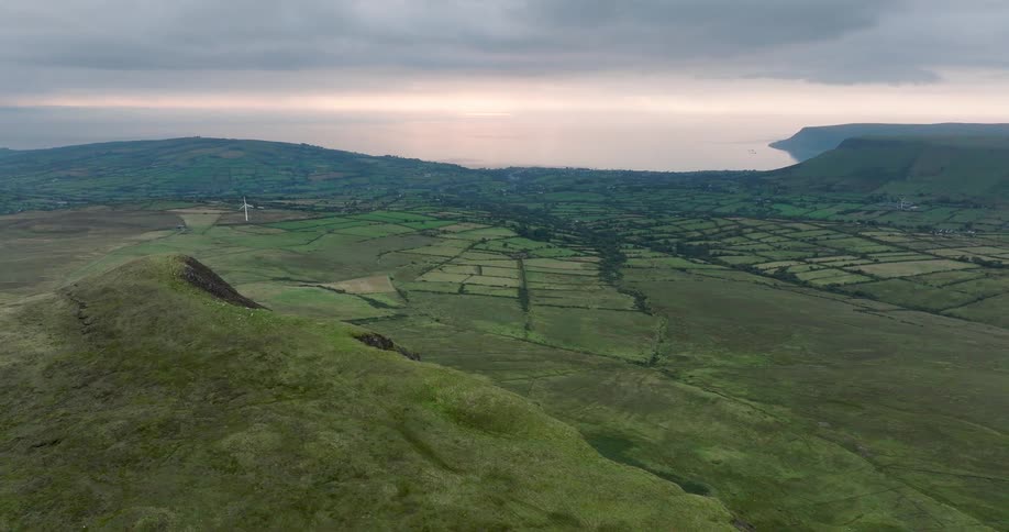 An aerial view pushes forward over the lush green rolling hills of the Irish countryside towards the distant coastline under a cloudy sky