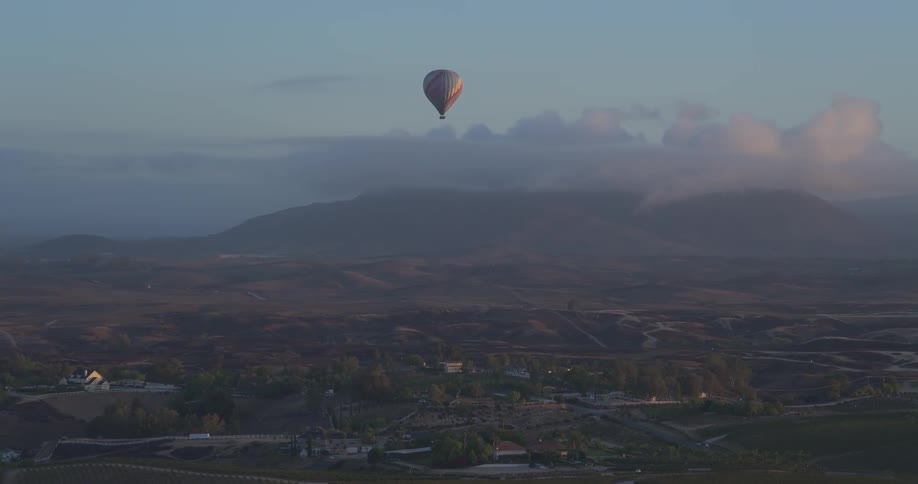 A lone hot air balloon soars gracefully over a vast, hazy valley with mountains and low clouds at sunrise