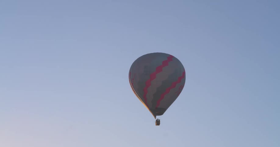 A solitary hot air balloon with pink and gray stripes floats gracefully across a serene morning sky at sunrise