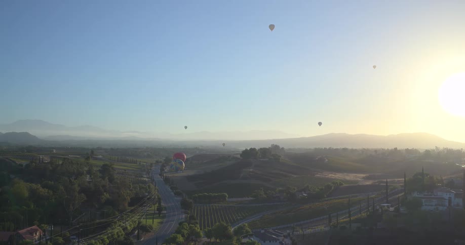 Slow aerial shot of hot air balloons peacefully drifting over a picturesque valley with vineyards at sunrise