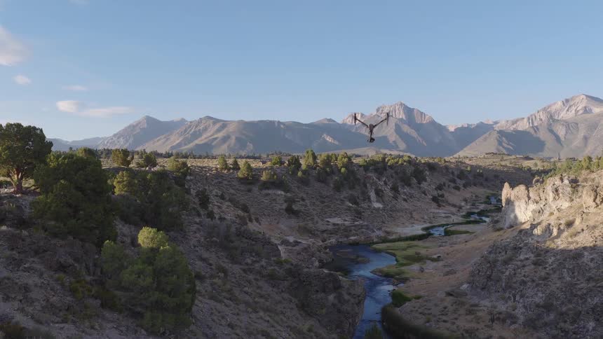 A professional filming drone hovers over a scenic river valley with a majestic mountain range in the background