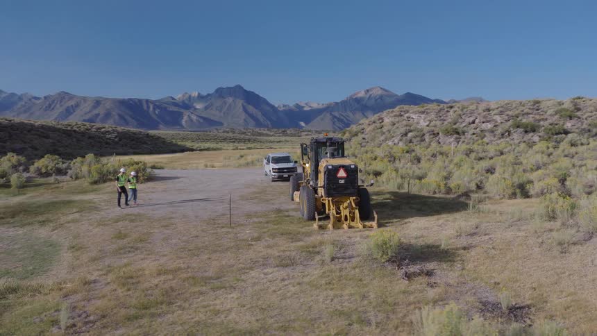 Two surveyors inspect a rural construction site with a motor grader parked nearby against a stunning mountain backdrop