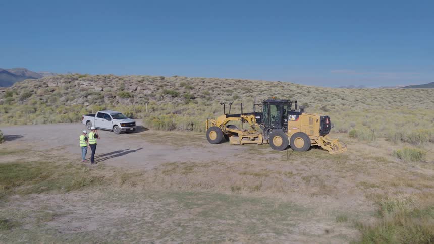 An aerial view reveals two construction workers standing next to a road grader at a rural worksite with majestic mountains in the background