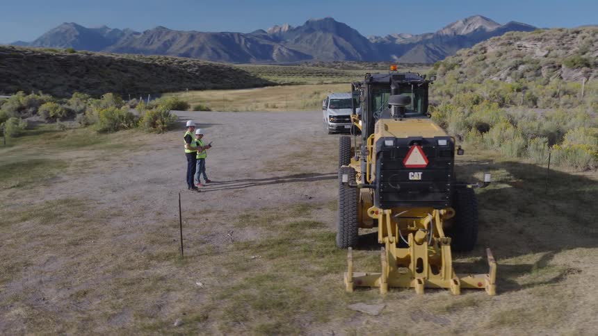 An aerial shot slowly pulls back from two construction workers standing by a road grader at a rural worksite with mountains in the distance
