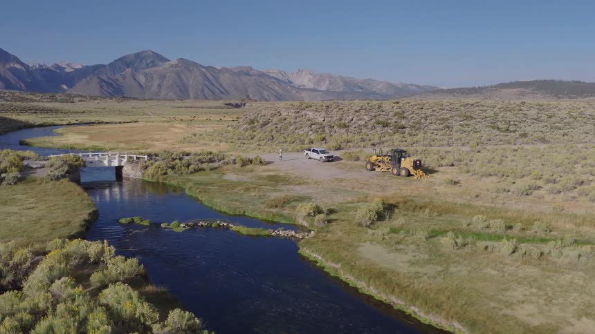 Aerial view of a worker surveying a construction site with a road grader next to a river in a vast mountain landscape