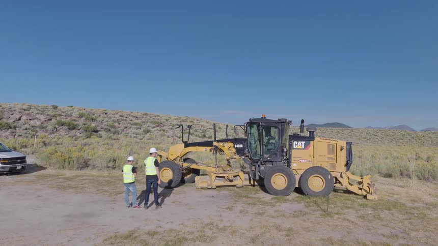 Aerial view of two construction workers in safety vests and hard hats discussing a project next to a motor grader on a rural job site