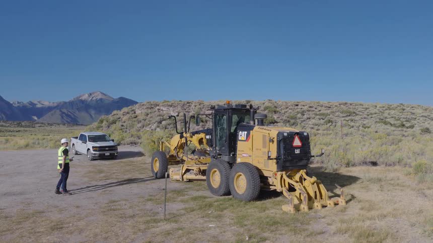 Aerial shot reveals two construction workers operating a drone next to a large motor grader in a vast, open landscape with mountains in the background