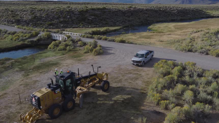 An aerial view pulls back from two construction workers talking next to a road grader on a rural dirt road with mountains in the background