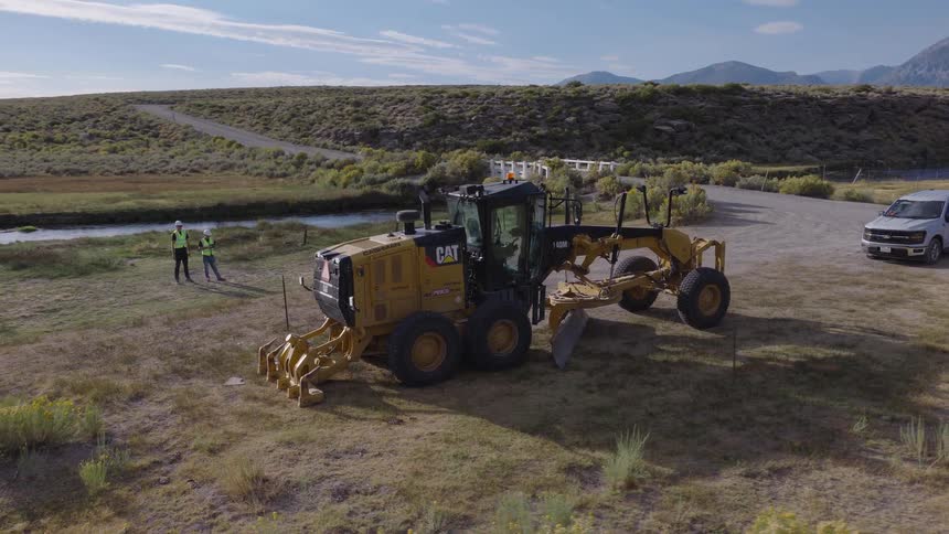 An aerial view captures construction workers operating a motor grader in a vast, arid landscape with mountains in the background