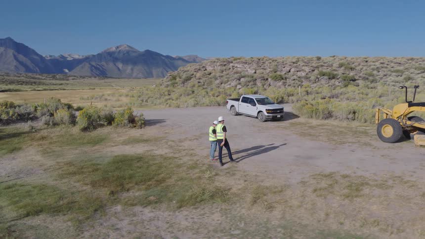 Aerial view of two engineers operating a drone at a rural construction site with a road grader and mountains in the background