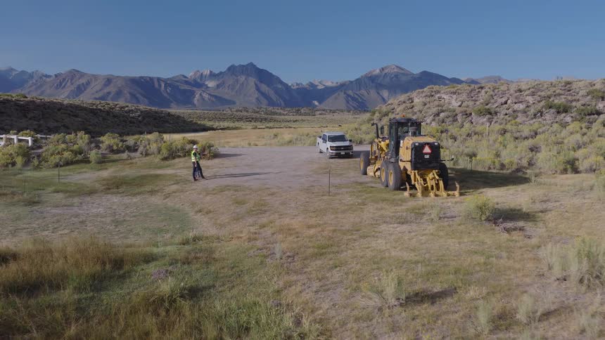 Aerial footage shows a surveyor operating a drone at a construction site with a motor grader, set against a beautiful mountain landscape