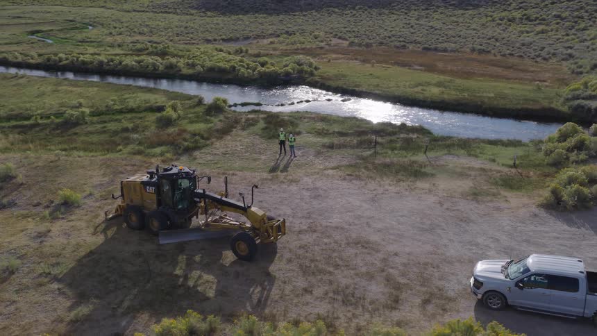 An aerial shot slowly rises above two surveyors and a motor grader parked by a winding river in a vast, open landscape