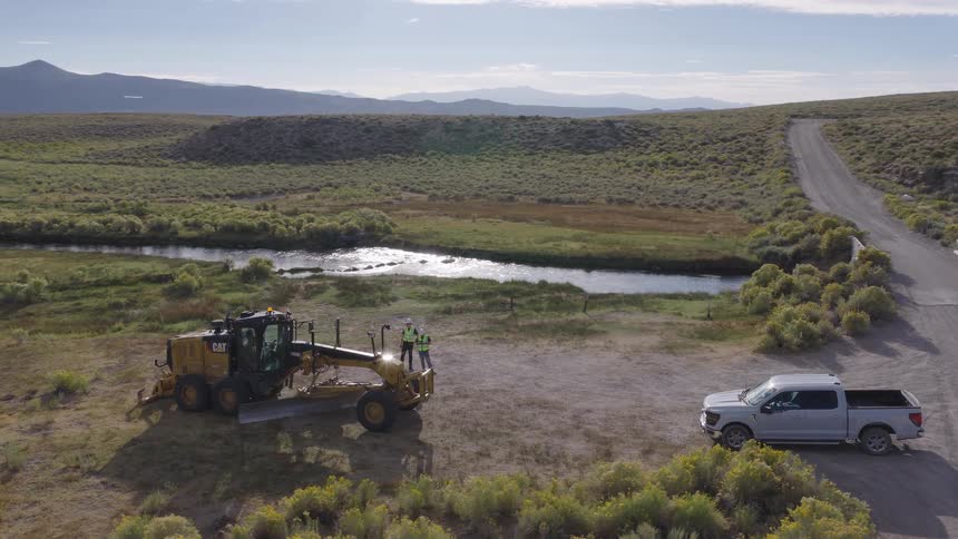 Aerial view of two construction workers standing by a CAT motor grader on a rural road job site next to a winding river
