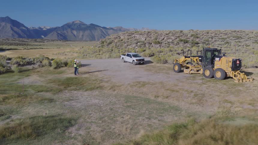 Aerial pullback shot reveals a construction worker operating a drone next to a motor grader and truck in a scenic mountain valley with a river and bridge